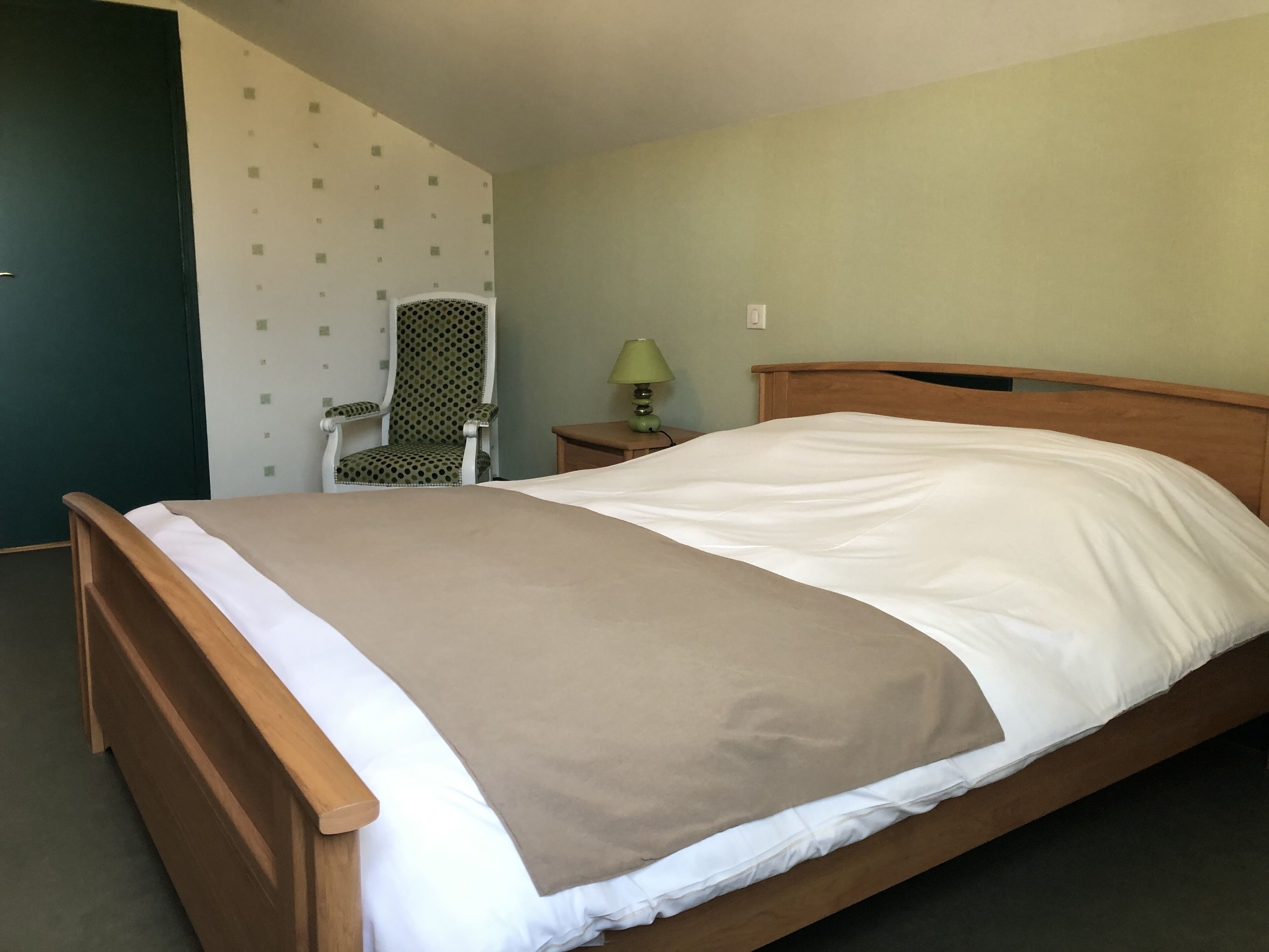 A single bed with a wooden frame and white bedding, a chair with a polka dot pattern, a small table with a lamp, and a wall with a simple pattern.