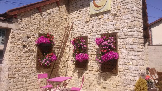 A stone building with a sign reading "La Colombelle Chambres Châteaux". There are two windows with pink flowers, a wooden ladder, and a table with chairs outside. - Maison d'hotes La Colombelle (Colombé-le-Sec)