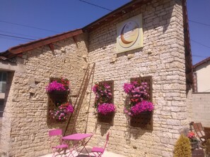 A stone building with a sign reading "La Colombelle Chambres Châteaux". There are two windows with pink flowers, a wooden ladder, and a table with chairs outside. - Maison d'hotes La Colombelle (Colombé-le-Sec)