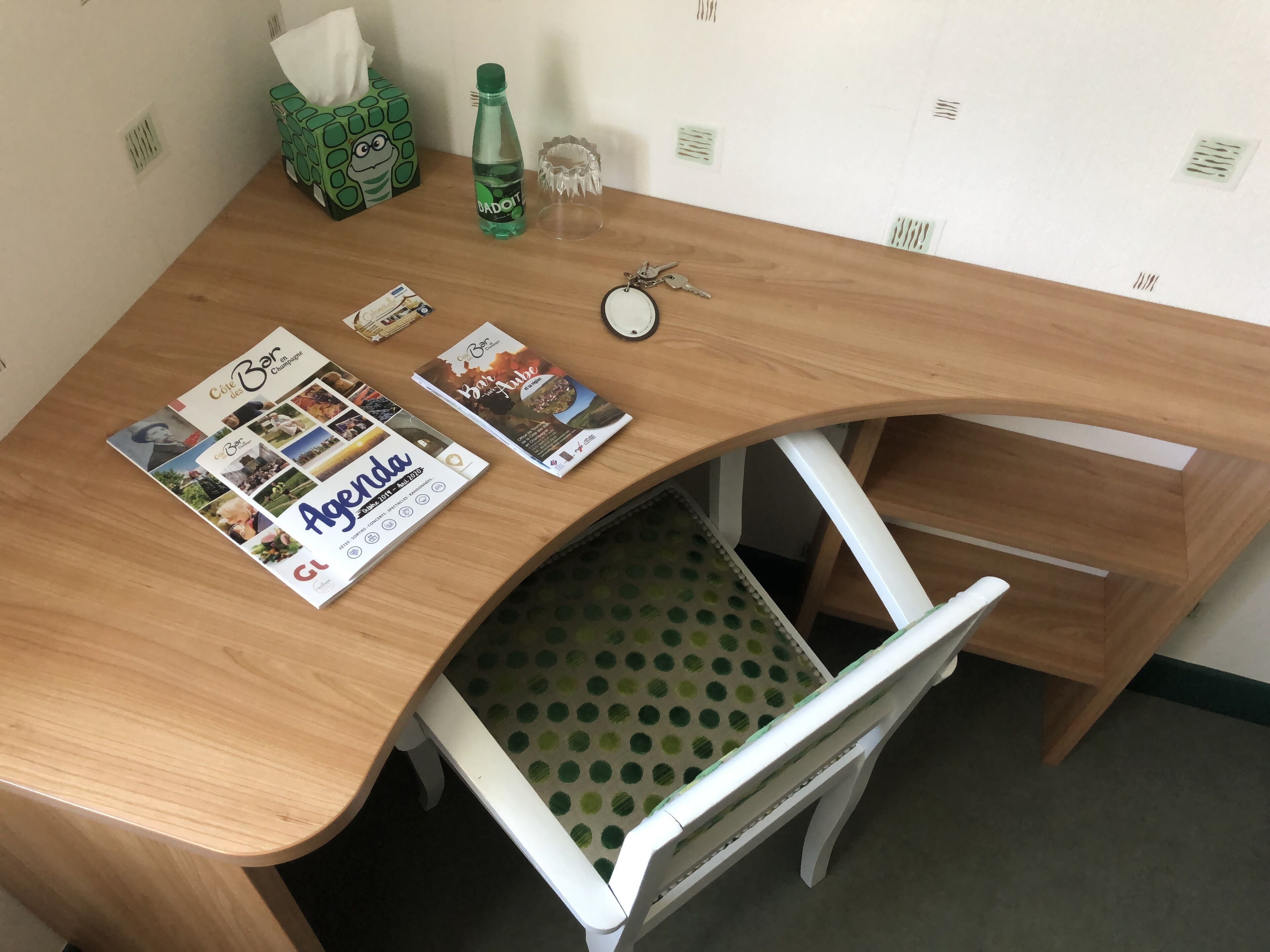 Un bureau en bois surmonté d’une nappe à pois, une boîte de mouchoirs, une bouteille, un trousseau de clés et un magazine.