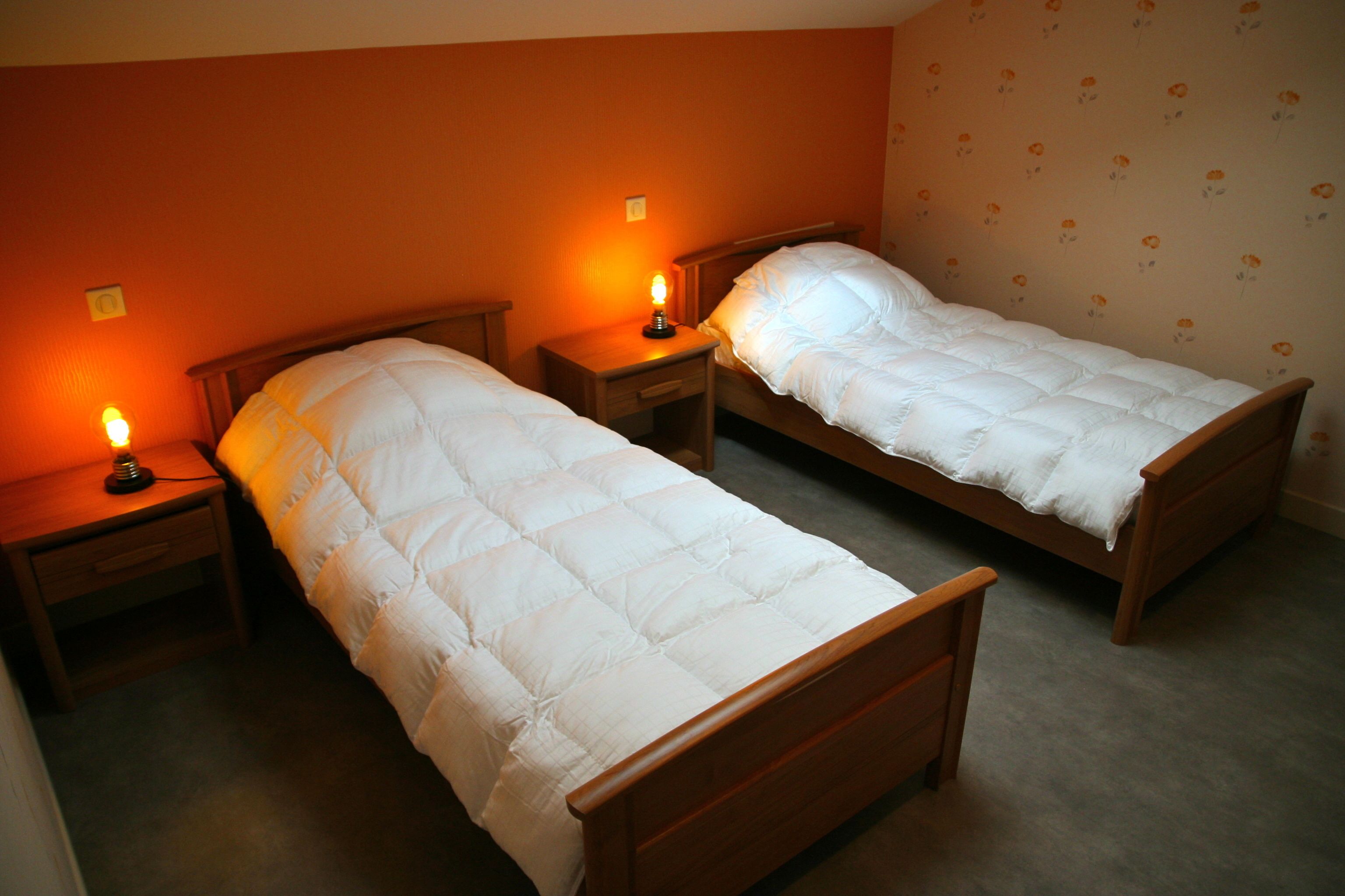 Deux lits simples avec des cadres en bois et du linge de lit blanc, dans une chambre aux murs orange et au papier peint à motifs floraux.