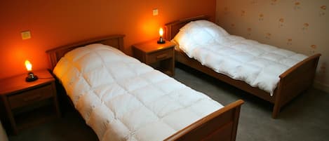 Deux lits simples avec des cadres en bois et du linge de lit blanc, dans une chambre aux murs orange et au papier peint à motifs floraux.