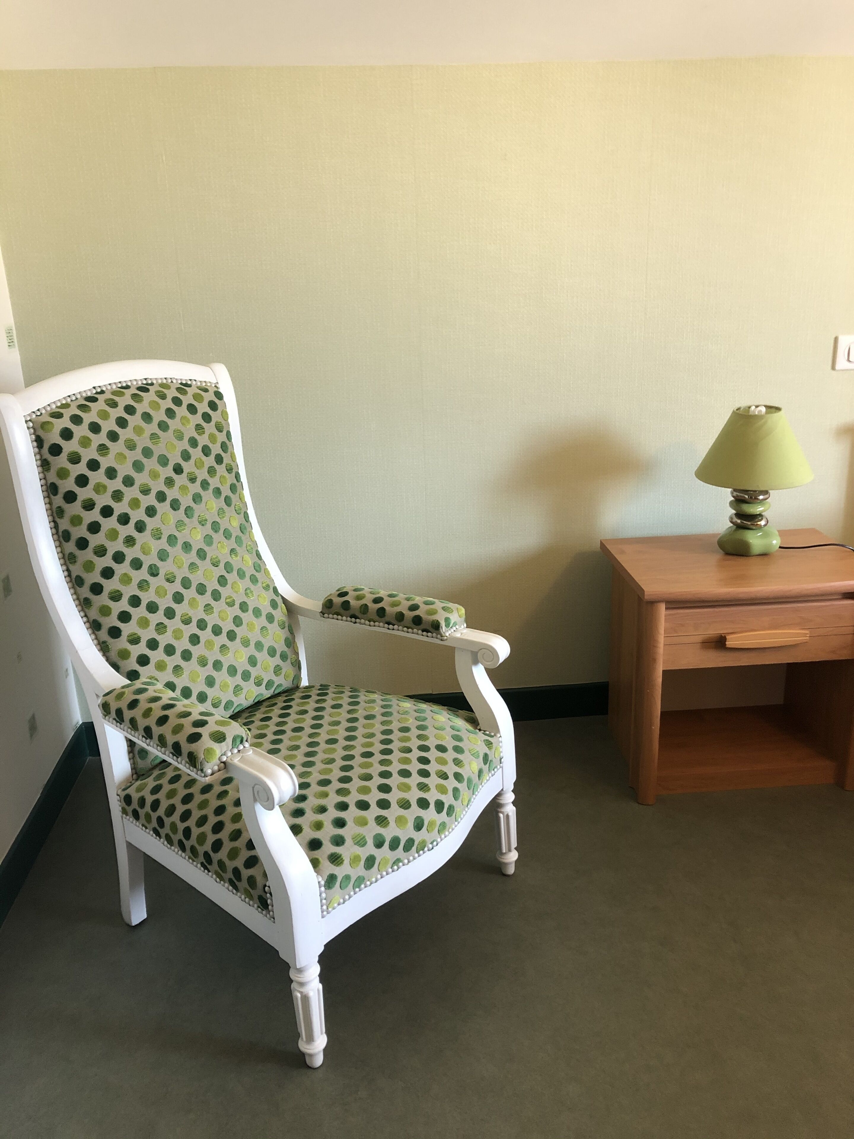 Una habitación con un sillón tapizado de lunares verdes, una mesita de madera con lámpara y una pared lisa.