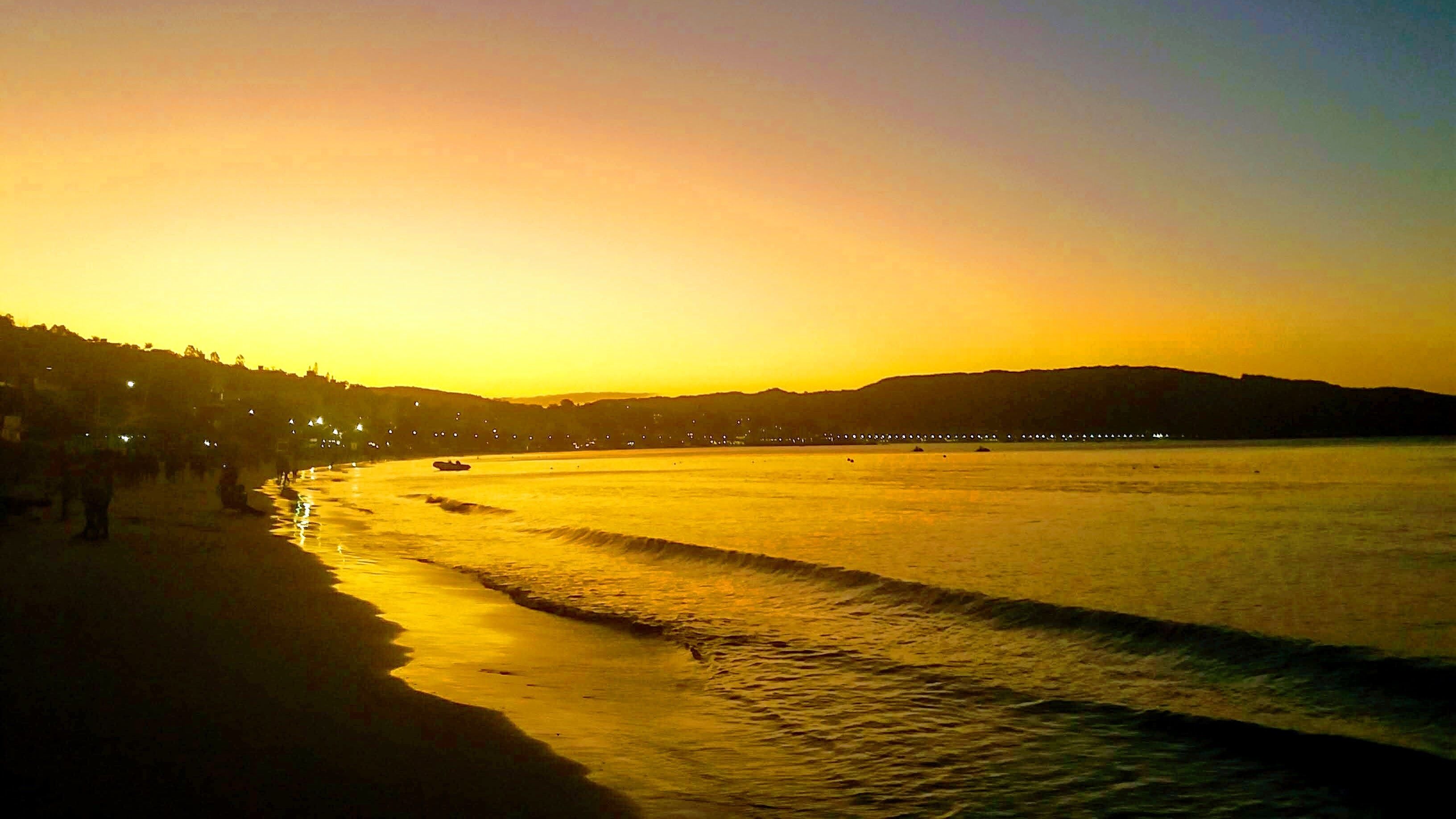A beach at sunset with calm waters and a distant shoreline.