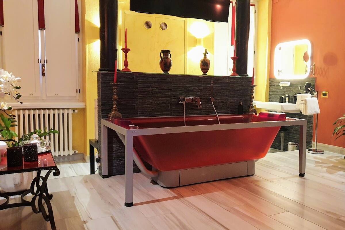 Une salle de bain moderne avec une baignoire rouge, un lavabo blanc et un miroir. On y trouve des vases décoratifs et une plante en pot.