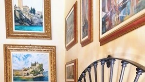 Ein Schlafzimmer mit einem Bett, einem Sessel und einer Person, die ein Buch liest. An der Wand hängen gerahmte Bilder.
