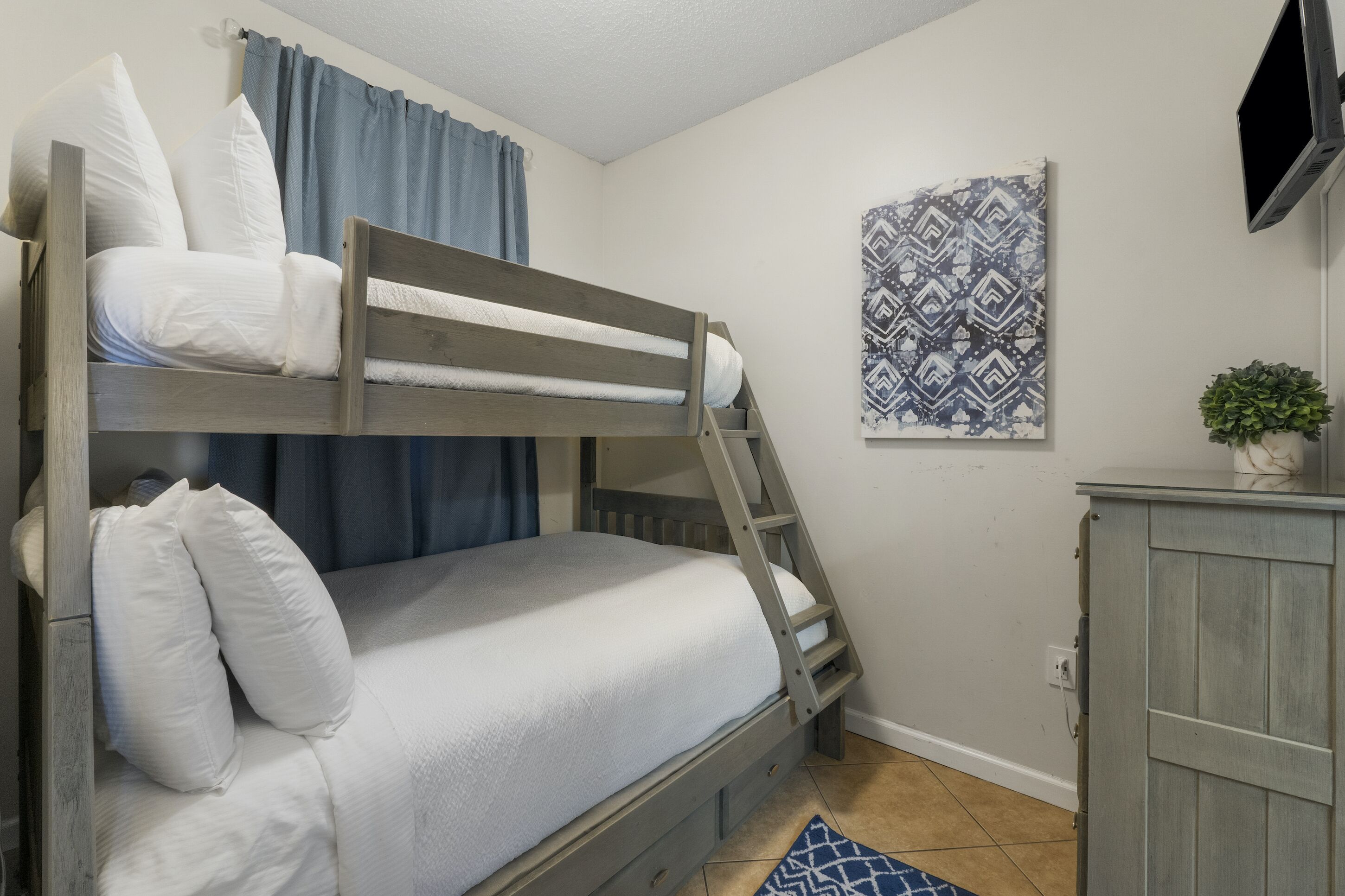Een stapelbed met witte beddengoed, een blauw gordijn, een wanddecoratie met patroon en een kleine plant op een kast.