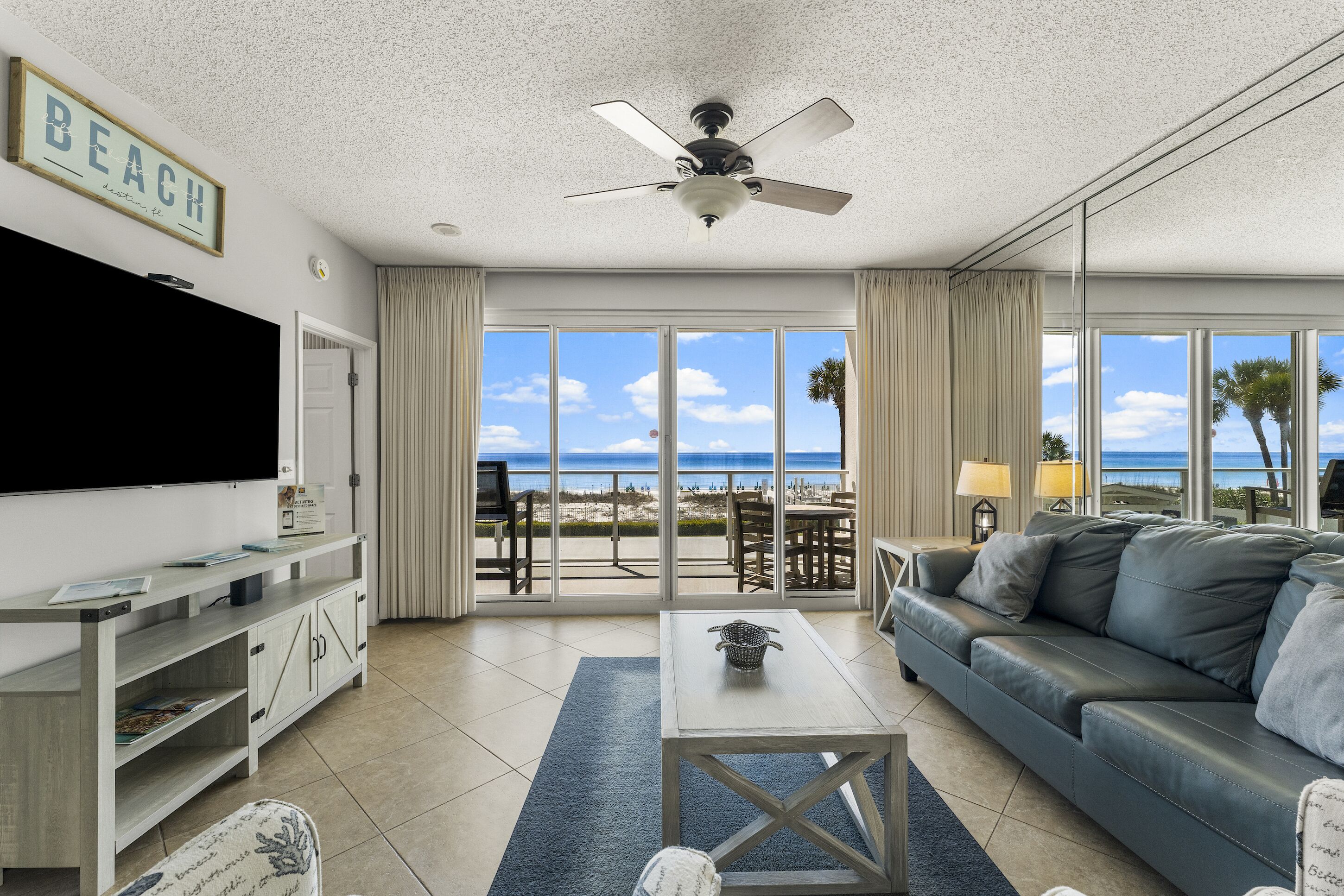 A living room with a large TV, a ceiling fan, a sofa, and a view of the beach through large windows.