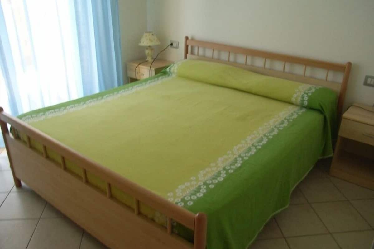 A bed with a wooden frame and green bedding, a nightstand with a lamp, and a window with curtains.