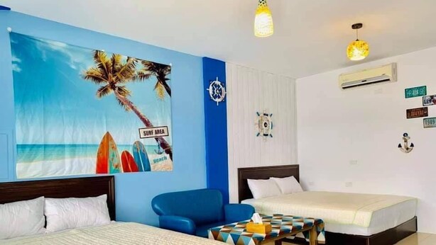 A hotel room with two beds, a blue armchair, a small table, and a beach-themed wall decoration. - Kenting Trail Villa (Hengchun)