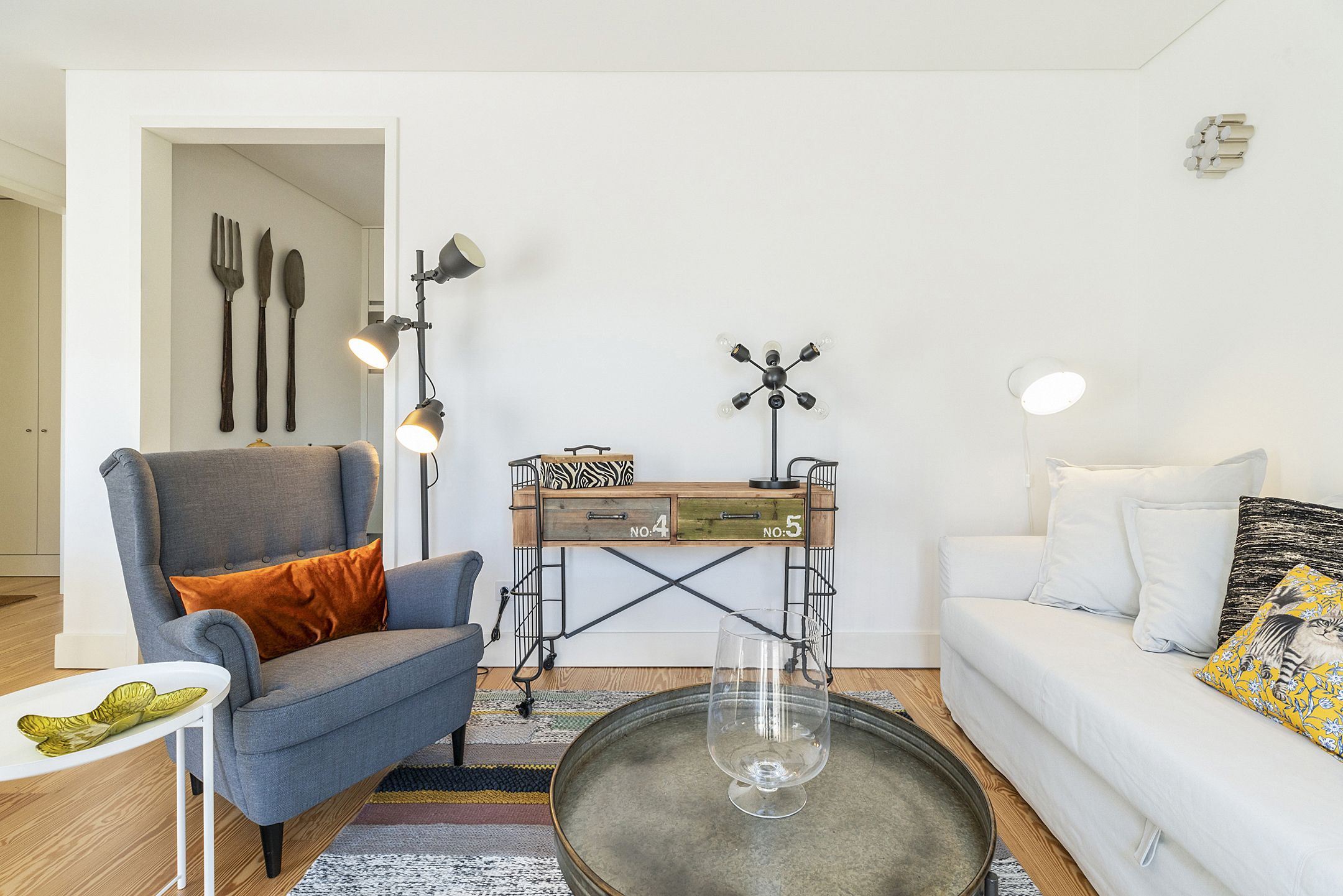 Una sala moderna con un sillón gris, un sofá blanco, una mesa de centro y una mesita auxiliar. Se observa un aparador de madera con cajones y una lámpara.