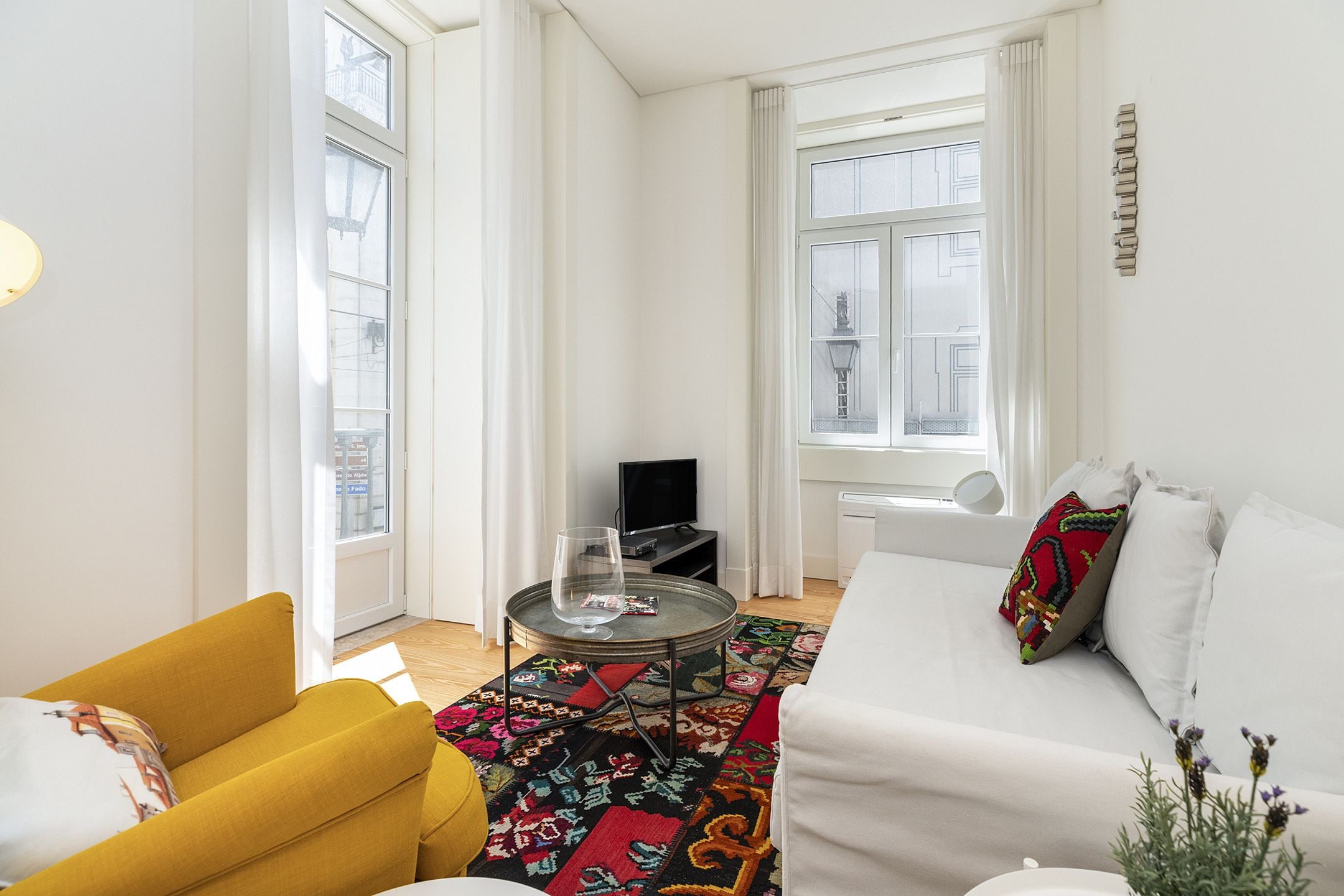 A modern living room with a yellow armchair, a white sofa, a round coffee table with a glass, and a colorful rug.