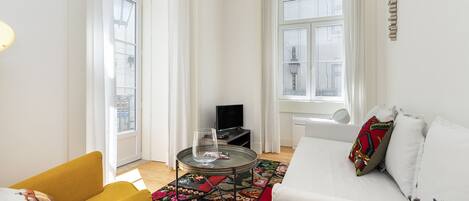 A modern living room with a yellow armchair, a white sofa, a round coffee table with a glass, and a colorful rug.