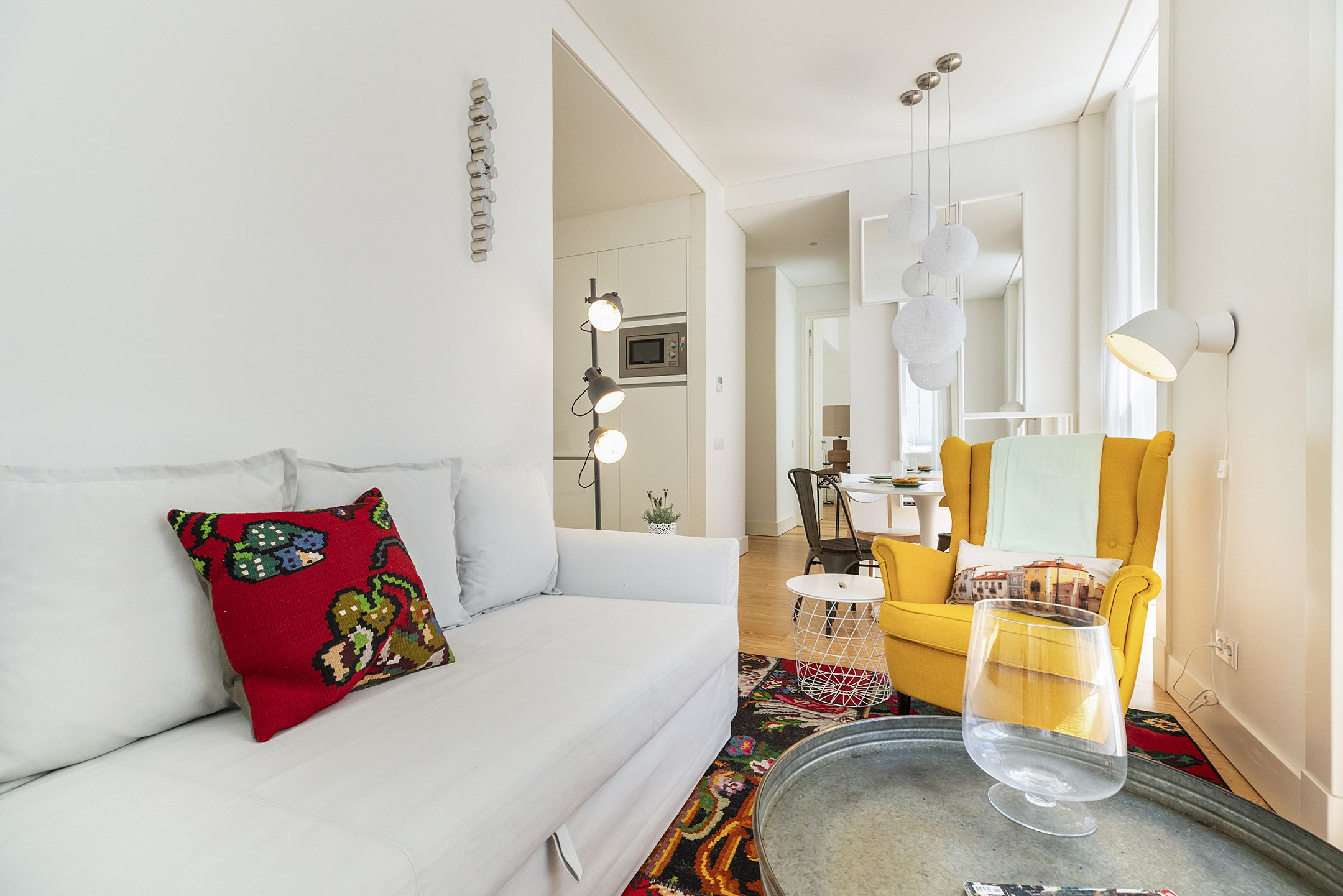 A modern living room with a white sofa, a colorful throw pillow, a yellow armchair, and a glass table with a wine glass.
