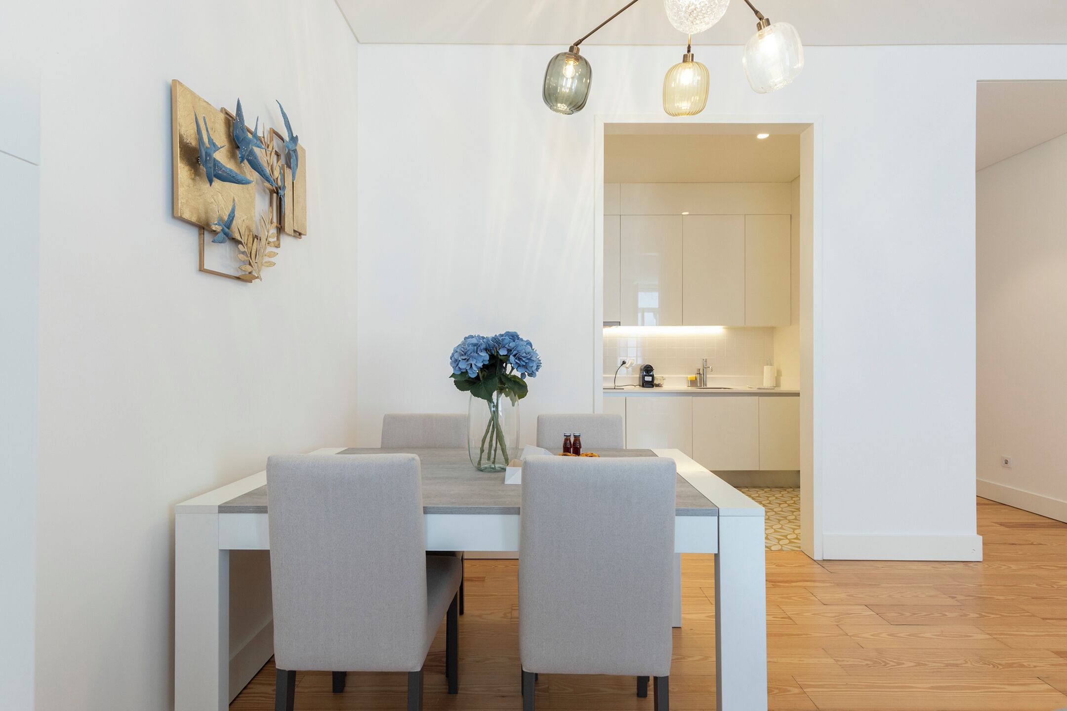 Un comedor moderno con una mesa y sillas blancas, un florero con flores y una cocina al fondo.