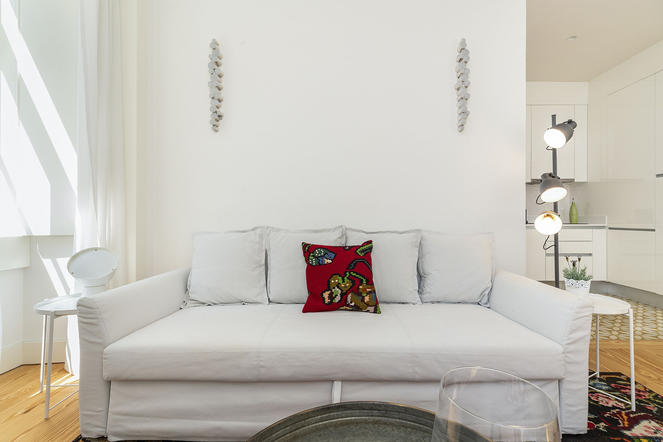 Een witte bank met een rood sierkussen, een klein wit bijzettafeltje en een vloerlamp in een moderne woonkamer, met een keuken op de achtergrond.