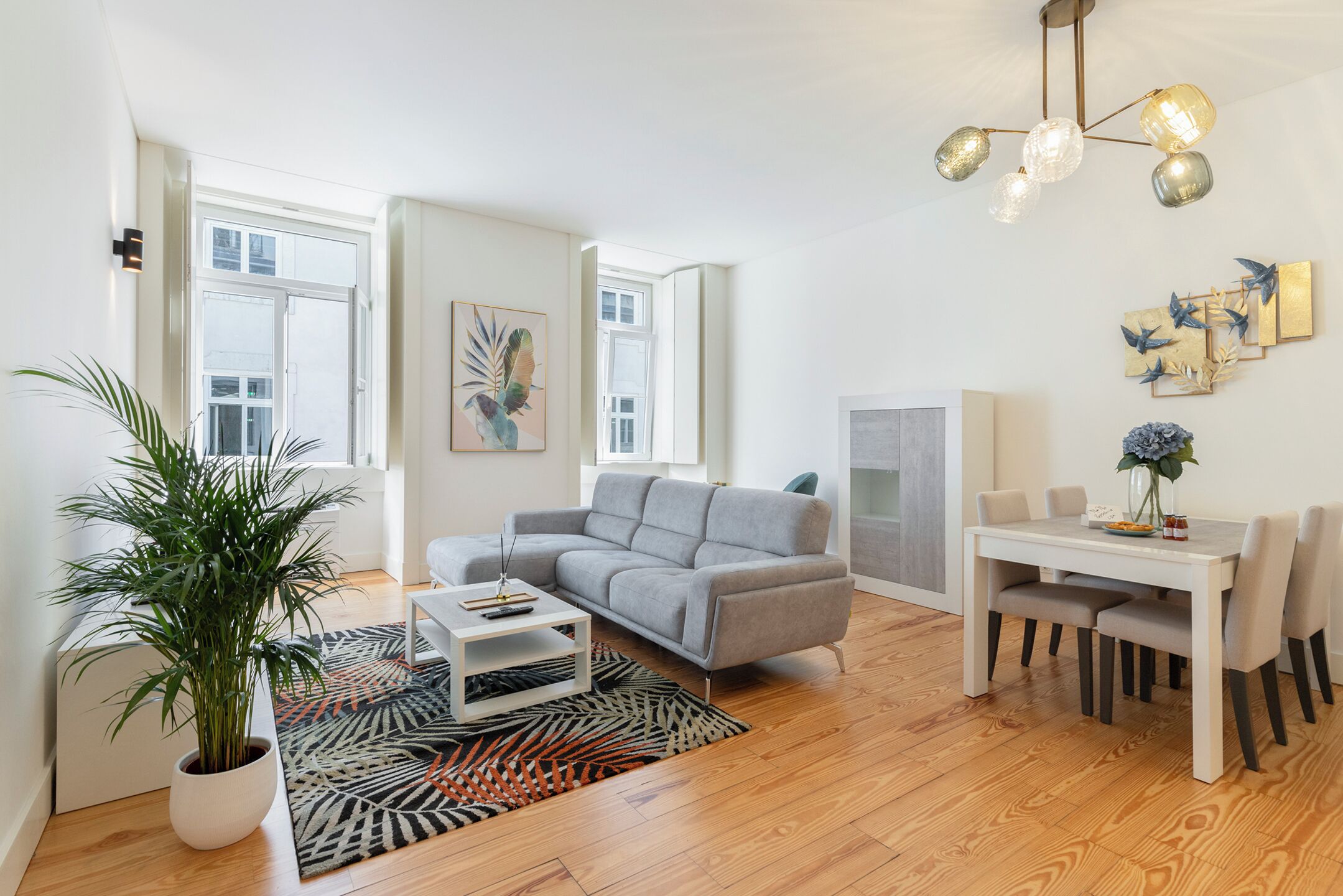 Una sala de estar moderna con un sofá gris, una mesa de comedor con sillas, una planta en maceta y una alfombra estampada.