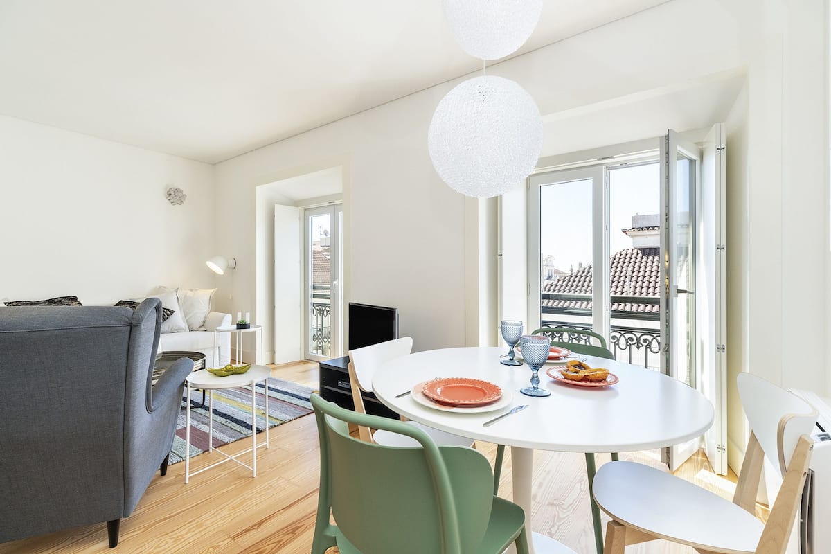A modern living room with a grey sofa, a white dining table with chairs, and a balcony with a view of buildings.
