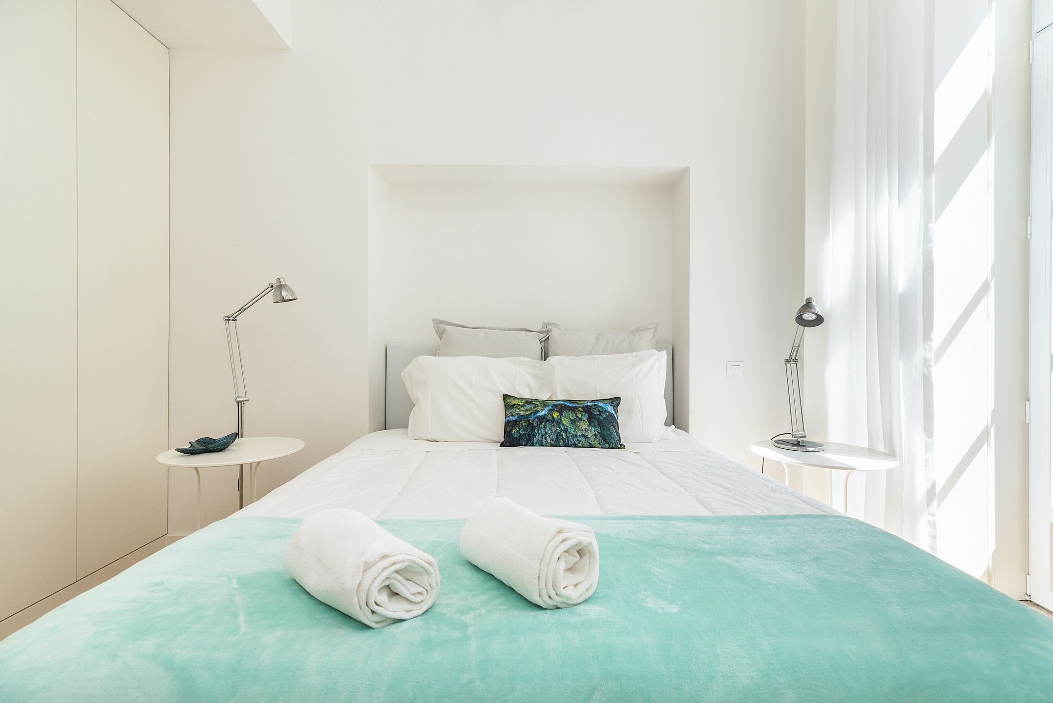 Habitación de hotel con cama, dos mesitas de noche, ventana con cortinas y una lámpara fijada a la pared.