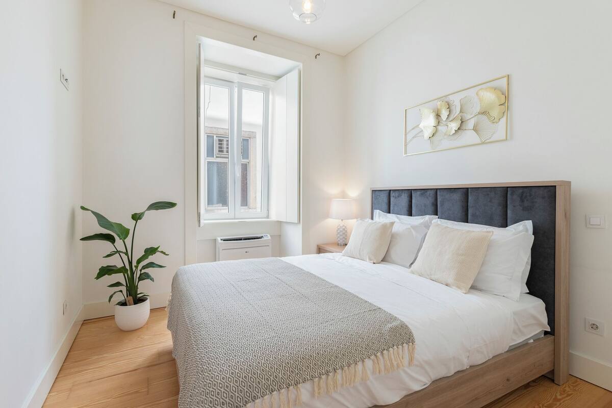 A bedroom with a large bed, a bedside table, a lamp, a window, and a potted plant.