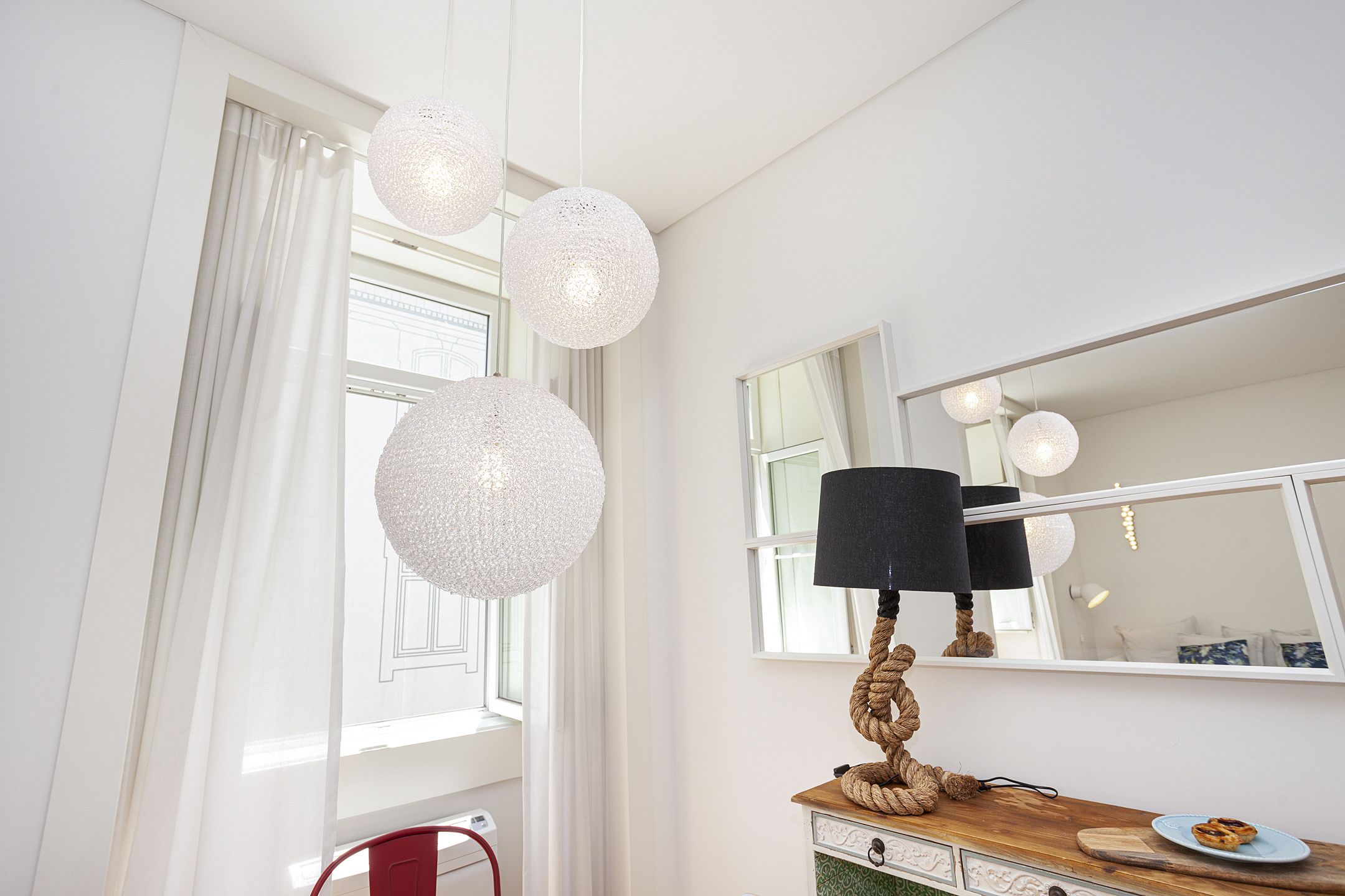 A room with a large window, white curtains, a wooden dresser, a mirror, and a round table with a plate.
