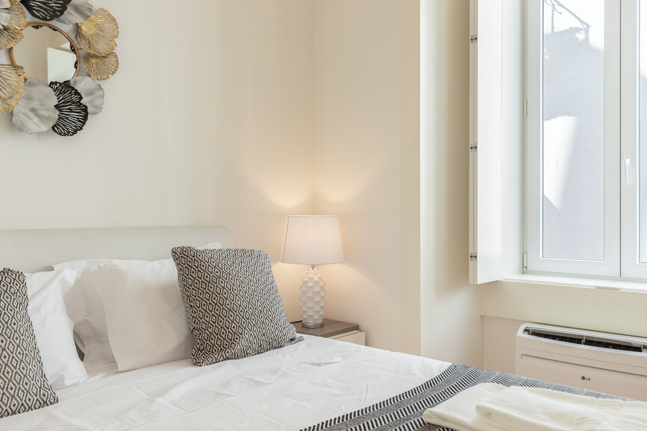 A neatly made bed with patterned pillows, a bedside lamp, and a window with blinds.