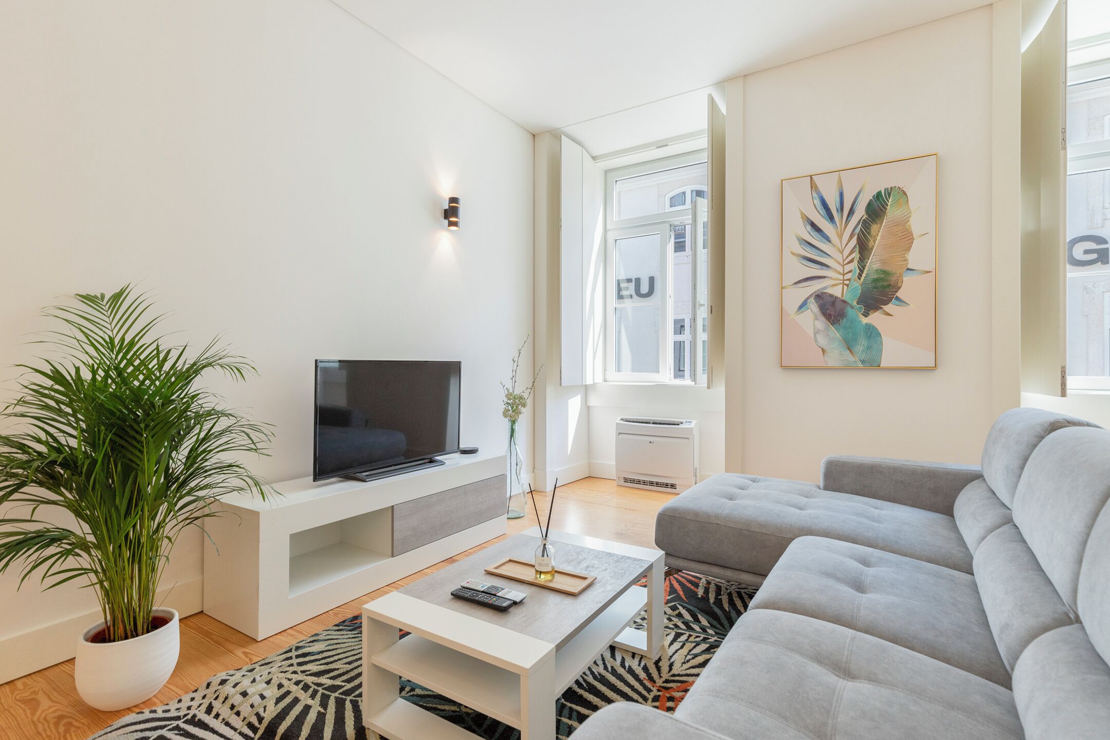 Ett modernt vardagsrum med en platt-TV, en grå soffa, ett vitt soffbord och en stor krukväxt.
