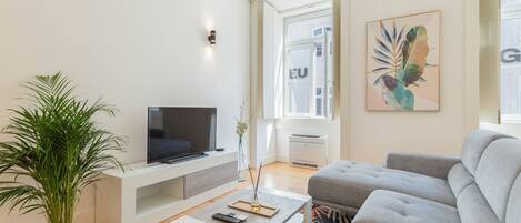 A modern living room with a flat-screen TV, a grey sofa, a white coffee table, and a large potted plant.