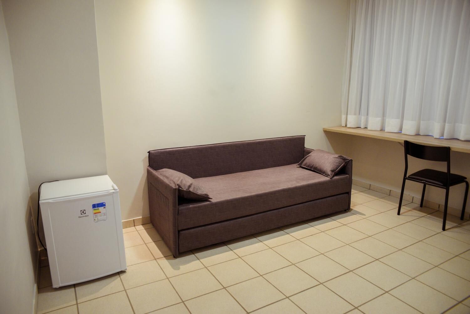 Una habitación pequeña y limpia con un sofá marrón, un refrigerador blanco compacto y una silla.