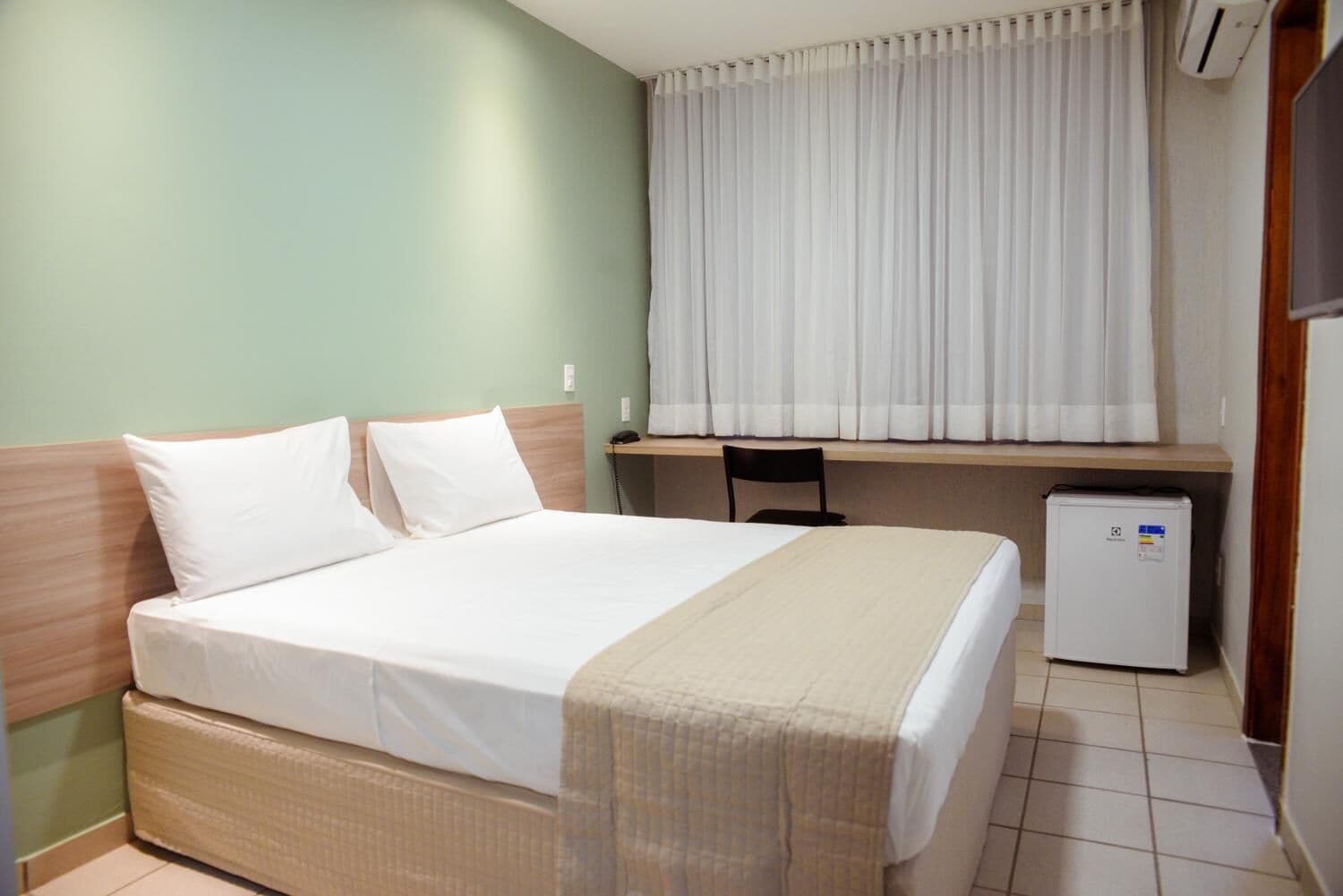 A hotel room with a bed, a desk, a chair, and a small refrigerator.