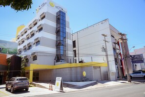 Un hotel moderno con fachada amarilla y blanca, varios balcones y un área de estacionamiento.