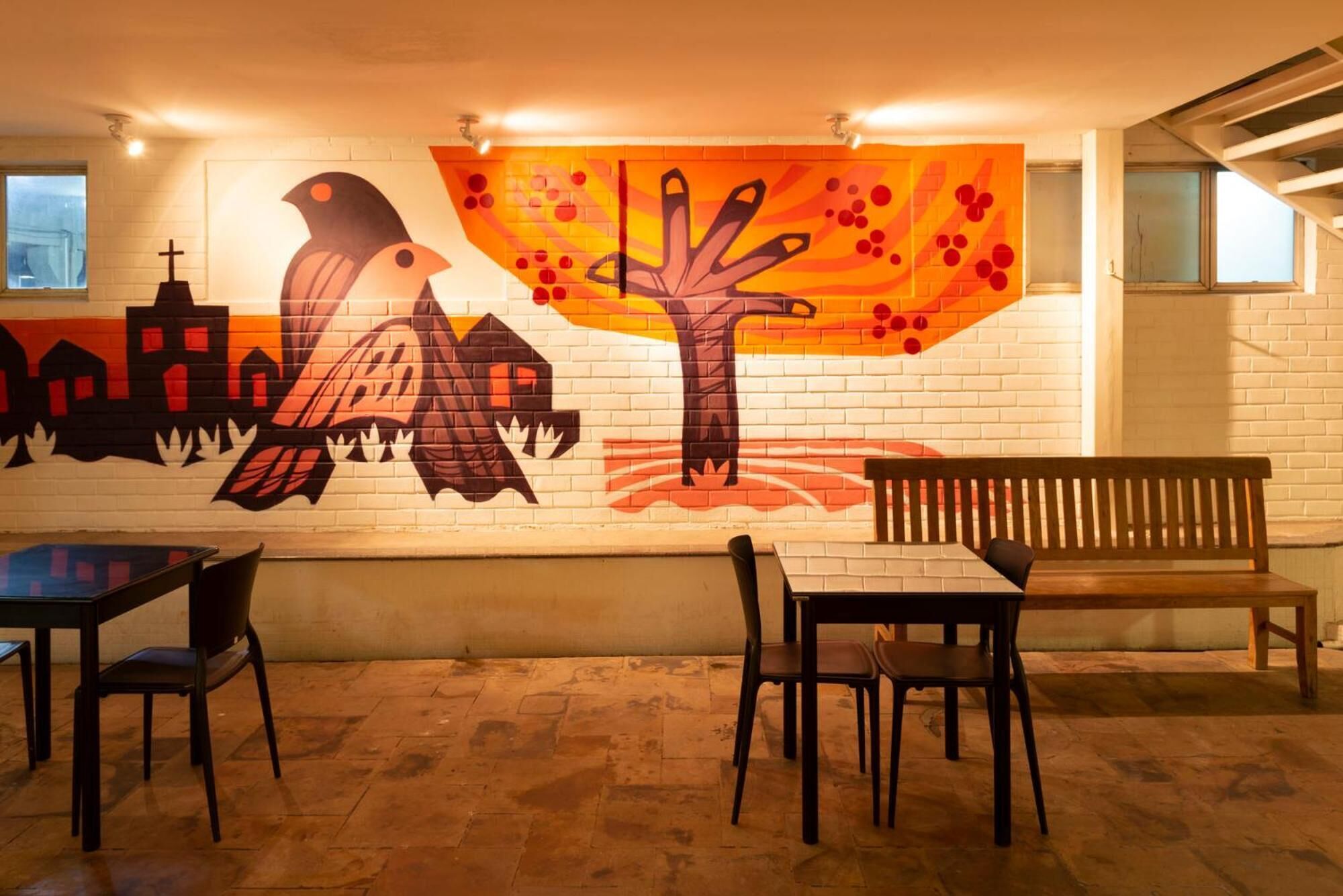 Un restaurante con un mural de un ave y un árbol en la pared, mesas y sillas de madera, y un banco.