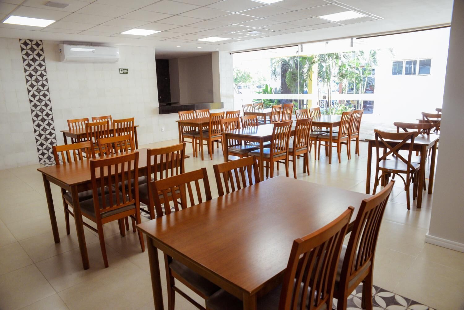 Un vaste espace dédié à la salle à manger, avec de nombreuses tables et chaises en bois, un grand téléviseur à écran plat et de grandes fenêtres offrant une vue sur les palmiers et les bâtiments.