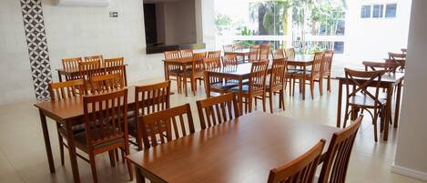 Un vaste espace dédié à la salle à manger, avec de nombreuses tables et chaises en bois, un grand téléviseur à écran plat et de grandes fenêtres offrant une vue sur les palmiers et les bâtiments.