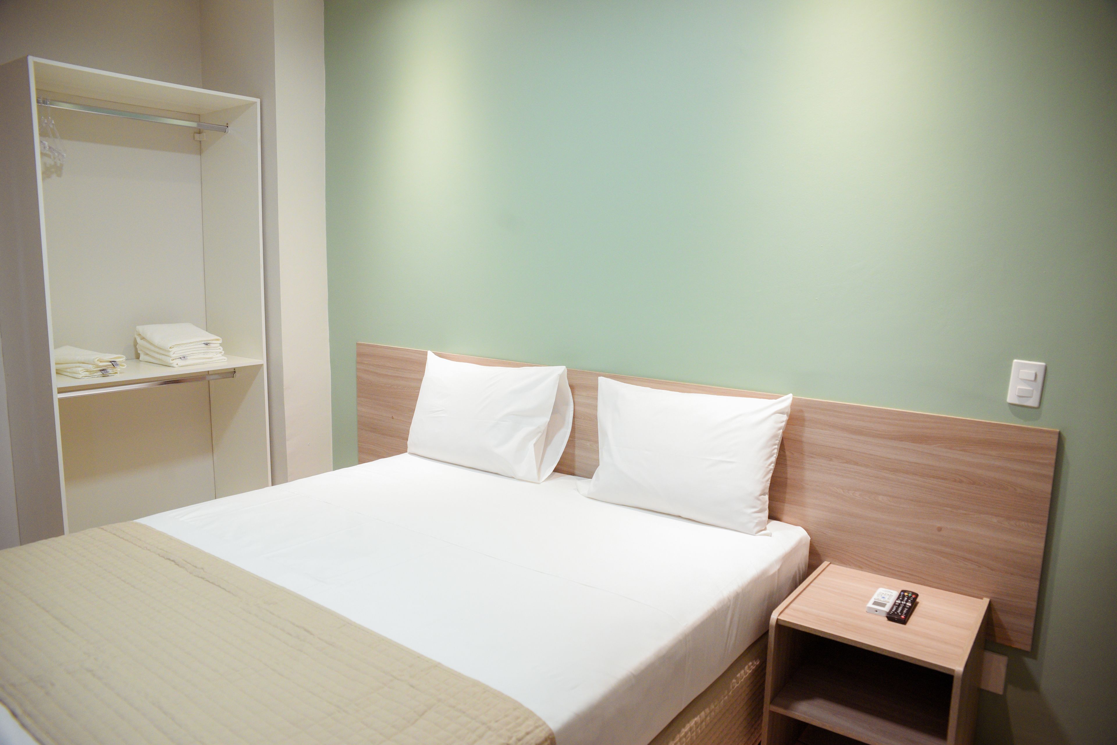A neatly made bed with white linens and pillows, a bedside table with a remote control, and a closet with folded towels.
