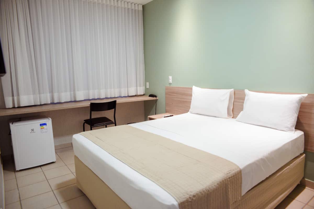 Ein Hotelzimmer mit Bett, Schreibtisch, Stuhl, kleinem Kühlschrank und einem Fenster mit Vorhängen.