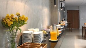 Un espace moderne dédié au petit-déjeuner dans un hôtel, avec un long comptoir, un buffet proposant divers plats et un vase rempli de fleurs jaunes.