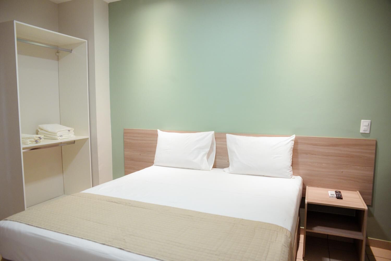 A neatly made bed with a wooden headboard, a bedside table with a remote control, and a wall-mounted shelf with folded towels.