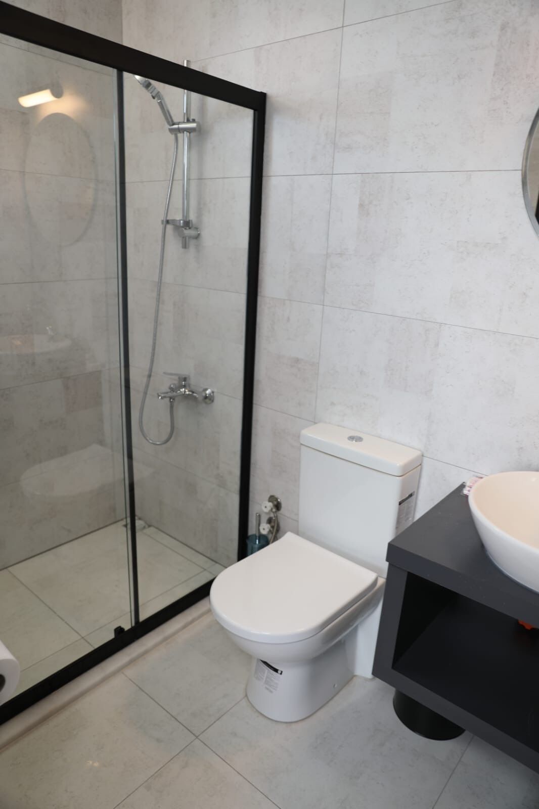 Un baño moderno con ducha de vidrio, inodoro blanco y tocador negro con lavabo redondo.