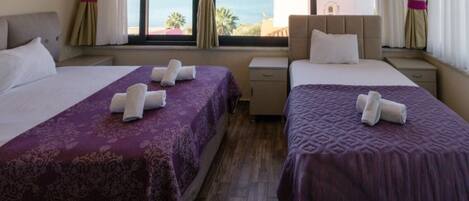 A hotel room with two beds, each with a purple bedspread and white towels. There is a window with curtains, a nightstand, and a view of the sea and a building.