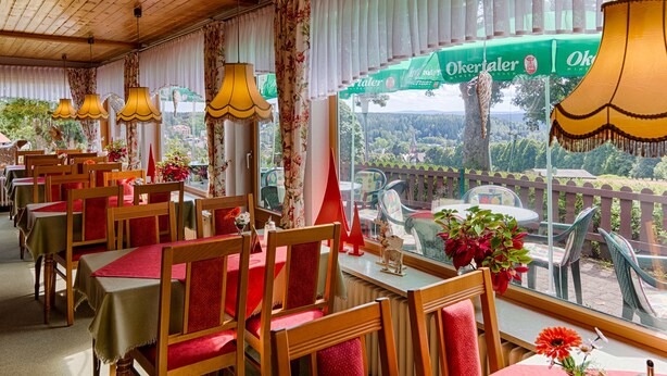 A restaurant with wooden tables and chairs, large windows with curtains, and outdoor seating visible through the windows. - Hotel & Café PANORAMA (Braunlage)