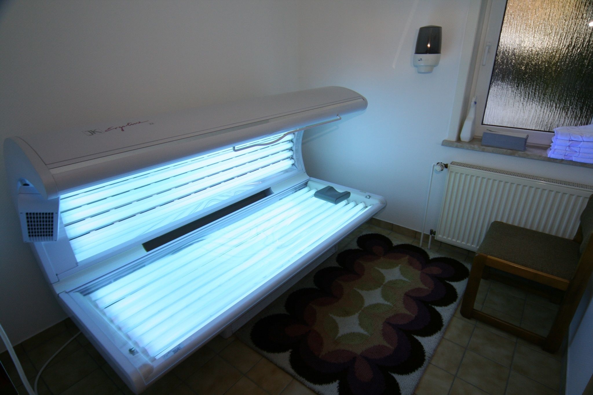 Una cama bronceadora en una habitación con un radiador, una mesita pequeña y una alfombra estampada.