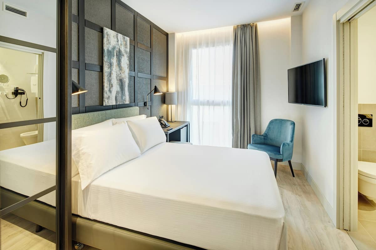 Ein Hotelzimmer mit einem großen Bett, einem Schreibtisch mit Lampe, einem Sessel, einem Fernseher und einem Badezimmer mit Toilette und Waschbecken.
