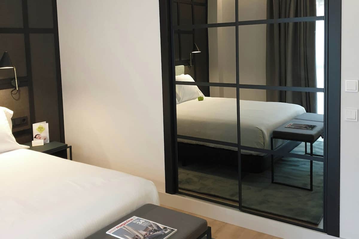 Moderni hotellihuone, jossa on sänky, yöpöytä ja suuri peili, joka heijastaa huonetta.
