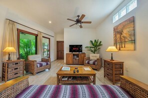 Ein Wohnzimmer mit Korbmöbeln, einem Deckenventilator, einem Fernseher und einem großen Fenster mit Vorhängen.