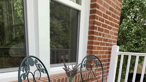 Dos sillas de metal con una mesa de mosaicos en el centro, ubicadas sobre una terraza de madera con un muro de ladrillo y una barandilla blanca al fondo.