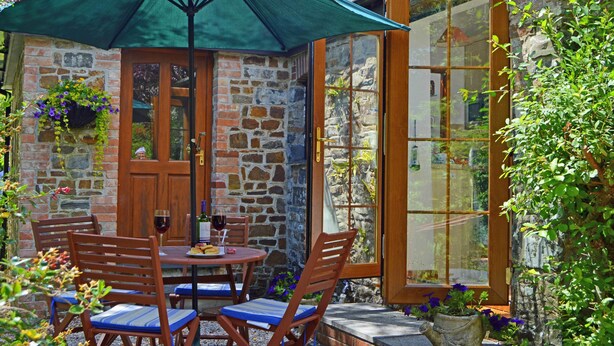 A stone building with a wooden door, a glass door, and a table with chairs under a green umbrella. - Shipload Cottage (Bideford)