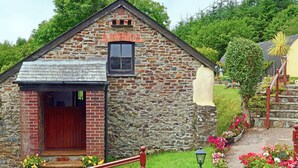 A stone building with a red door and a small window. There are flower beds with colorful flowers in front of the building. - Shipload Cottage (Bideford)