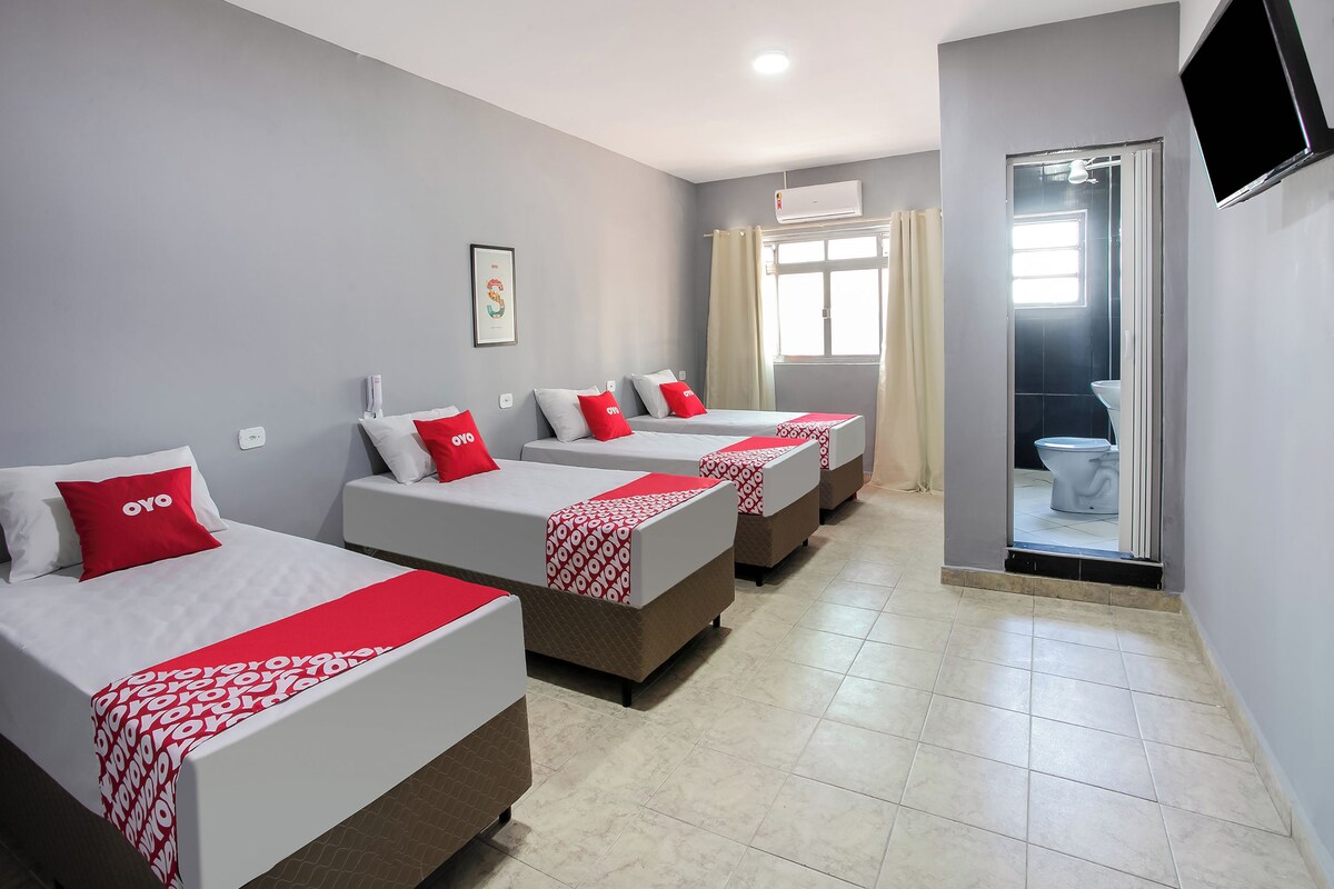 Une chambre d’hôtel avec trois lits, chacun recouvert d’une courtepointe rouge et blanche et agrémenté d’un oreiller orné du logo « oyo ». La chambre est équipée d’un luminaire au plafond, d’un climatiseur et d’un téléviseur fixé au mur.