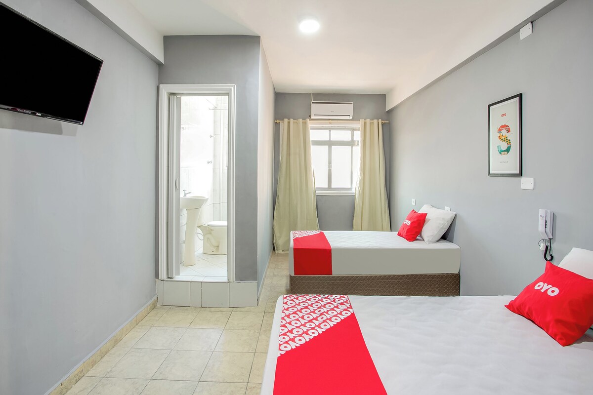 Une chambre d’hôtel avec un lit, une table de chevet, un téléviseur fixé au mur, une fenêtre avec des rideaux et une salle de bain visible à travers une porte ouverte.